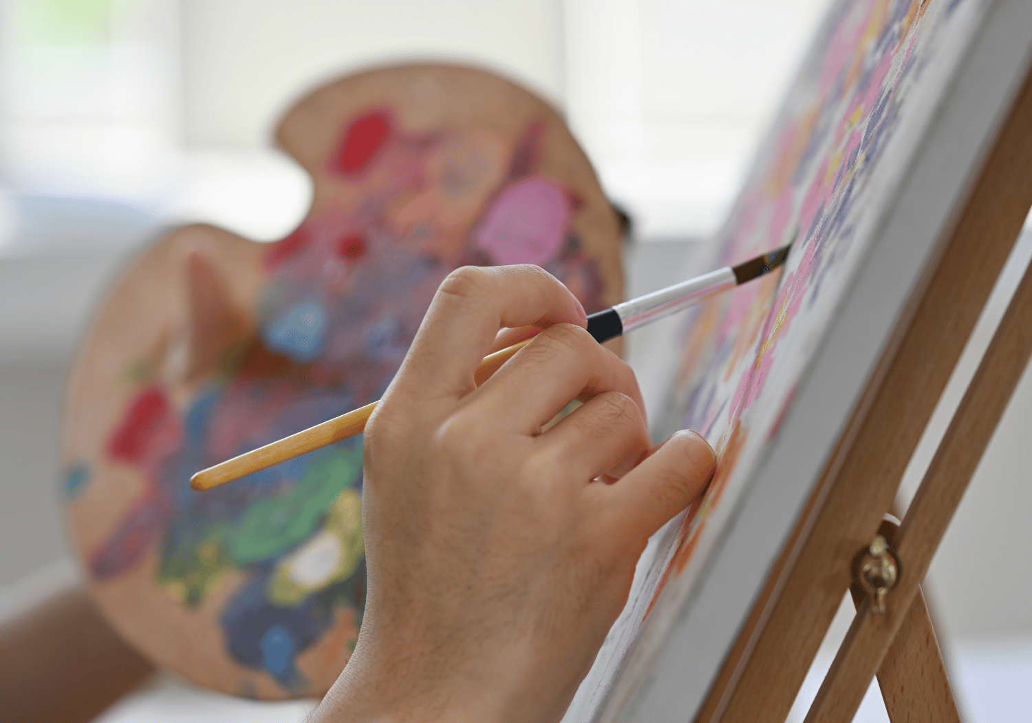Close-up of a hand holding a paintbrush and painting on a canvas set on an easel, with a colorful palette blurred in the background